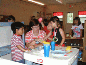 A teacher sits a low table assisting two four year olds with her back to the rest of the classroom.