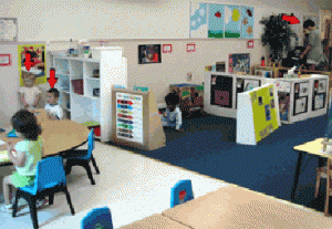 A teacher is on the far right of the classroom. Two children are behind tall shelves at the other side of the classroom