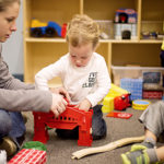 a preschooler sits on the floor beside a teacher assembling a train toy