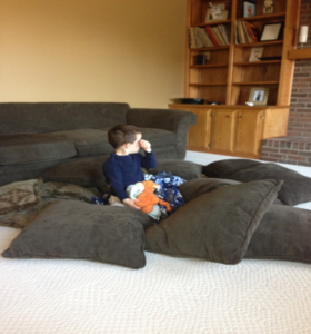 A boy sits on the floor sucking his thumb, surrounded by couch cushions