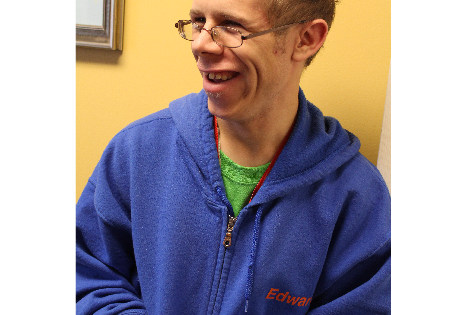 A young man stands smiling in front of a yellow wall, looking off camera.