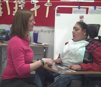A picture of a teacher sitting facing a student. The student is in a wheelchair, with a tray on the front. The teacher smiles as the student responds to her question.
