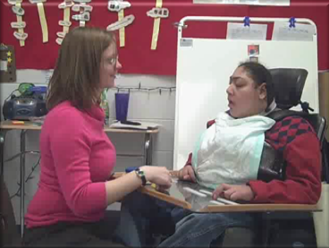 A picture of a teacher sitting facing a student. The student is in a wheelchair, with a tray on the front. The teacher smiles as the student responds to her question.