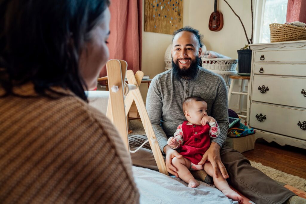 Man sitting in room in a house holding a small child in his lap.  He is smiling at someone across from him.