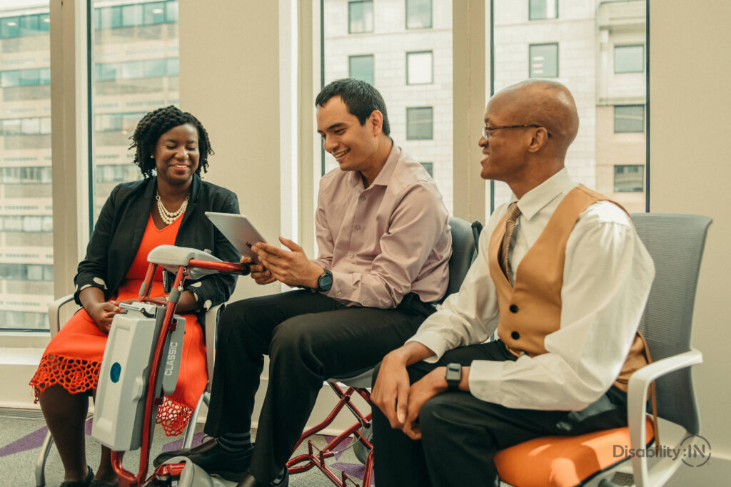 In this image, three professionals are having a conversation. They sit before a window overlooking a city. The woman on the far left is wearing an orange dress and black jacket. The man in the middle is holding a tablet and wearing a tan shirt and black slacks. The person to the right is looking away from the camera, and has a dress shirt, tie, and yellow vest on.