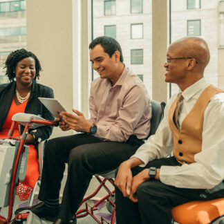 In this image, three professionals are having a conversation. They sit before a window overlooking a city. The woman on the far left is wearing an orange dress and black jacket. The man in the middle is holding a tablet and wearing a tan shirt and black slacks. The person to the right is looking away from the camera, and has a dress shirt, tie, and yellow vest on.