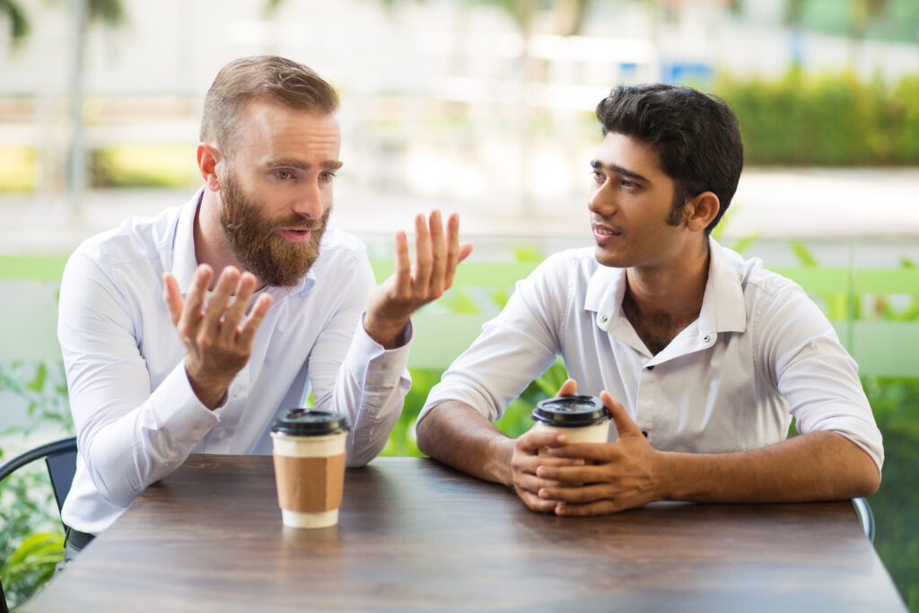 Two male friends drinking coffee and talking in outdoor cafe. People sitting at table with blurred view in background. Coffee break concept. Front view.