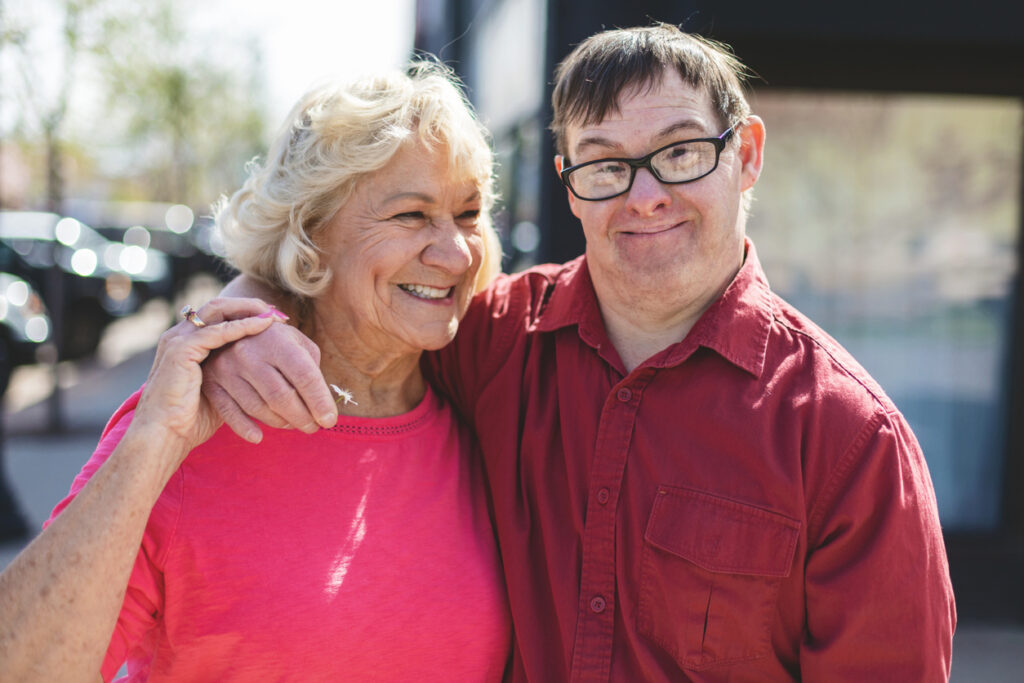 Adult Man Portraits with a Down Syndrome Outdoors hugging the Senior Female Guardian 