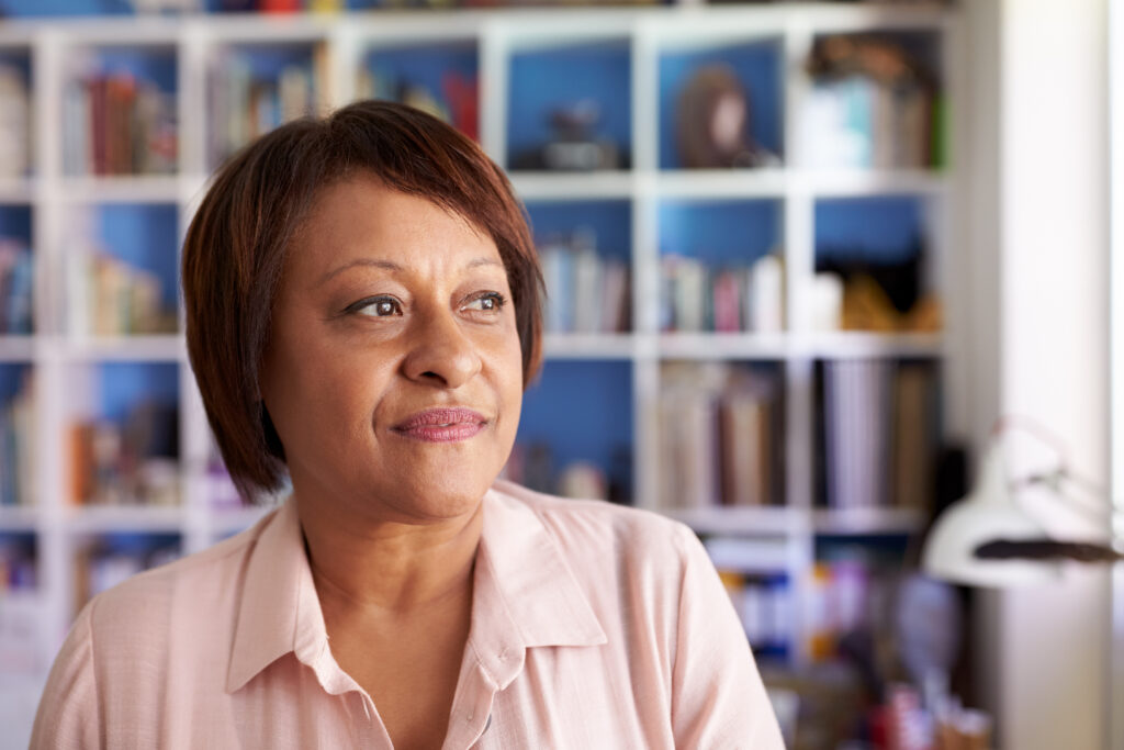 Middle aged african american woman sitting in front of a wall bookshelf looking off to the distance.