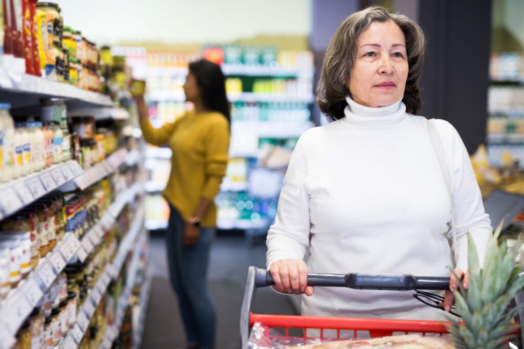 Older woman wearing a white shirt pushing s shopping cart in a grocery store.