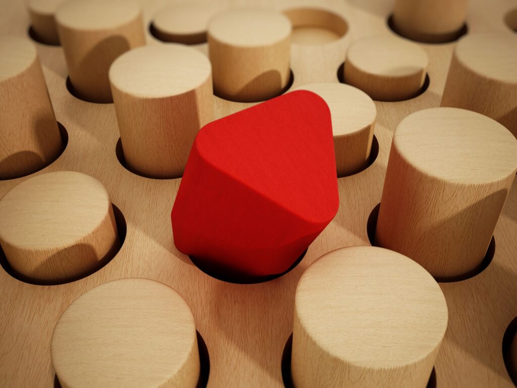 Brown wooden puzzle with several cylindrical pegs.  Red 3-D triangle sitting, not fitting, in a circular peg hole.
