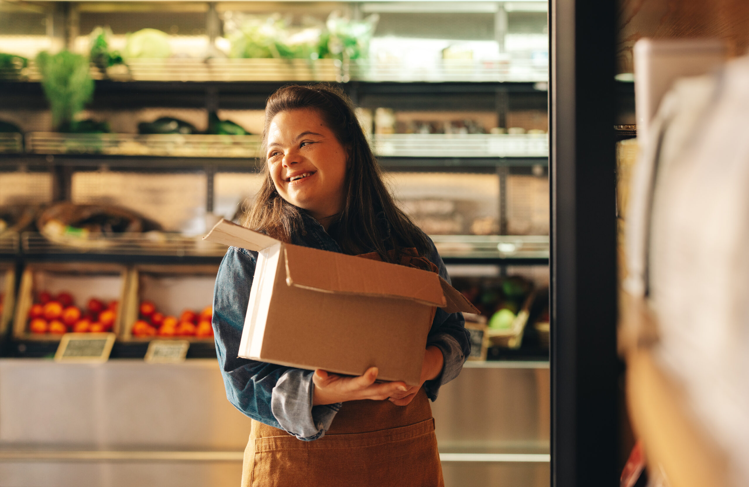 Woman with Down syndrome smiling happily while working as a shopkeeper in a grocery store. Empowered woman with an intellectual disability restocking food products in a supermarket.