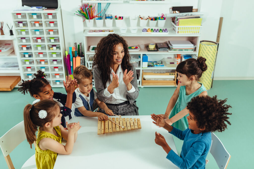 A classroom with children singing with their teacher at a table.
